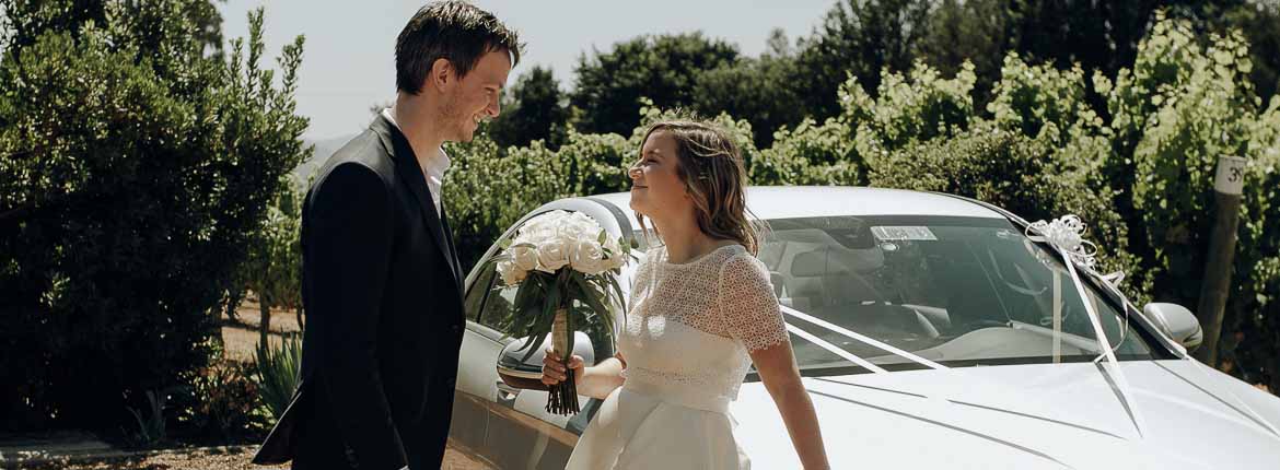 The bride and groom are in front of each other, if looking into the eyes, the bride holds the bouquet of roses and is leaning against a silver Mercedes.
