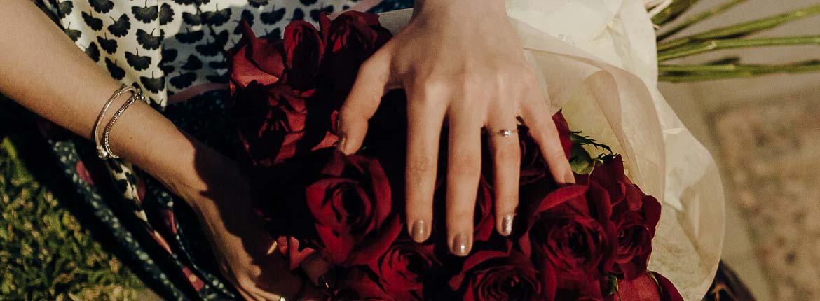 Mão da Tamires com aliança no dedo e mão está acima do ramalhete de rosas vermelhas.